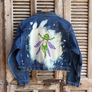 Op de afbeelding: Een blauwe denimjas met een wit gebleekt ontwerp op de rug. Het ontwerp toont een groene fee met paarse vleugels. De handtekening van de kunstenaar, "Lizzy Meadows", is zichtbaar onder de fee.