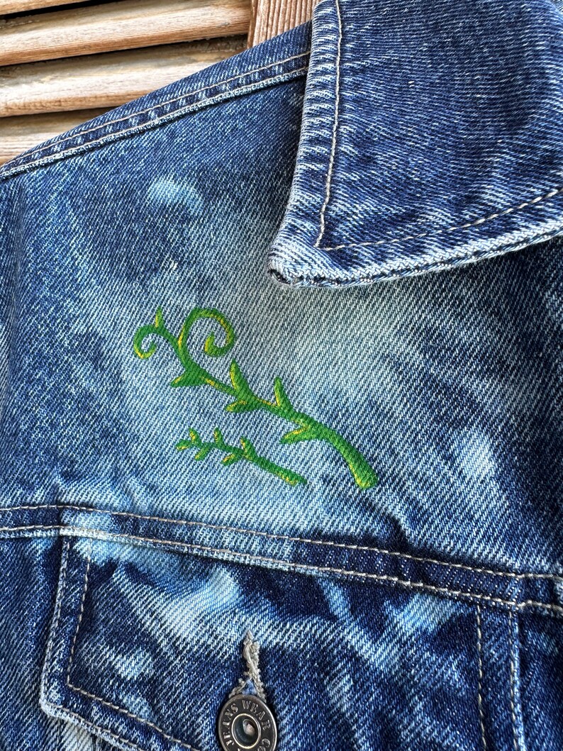 Pu&ograve; includere: Una giacca di jeans blu con un disegno di vite ricamato in verde sul lato sinistro del petto.
