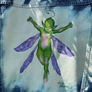 Pu&ograve; includere: Una giacca di jeans con un design dipinto a mano di una fata verde con ali viola. La fata sta volando con le braccia distese. La firma dell'artista, "Lizzy Houbiers", &egrave; nell'angolo in basso a destra del design.