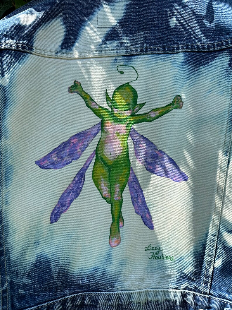 Pu&ograve; includere: Una giacca di jeans con un design dipinto a mano di una fata verde con ali viola. La fata sta volando con le braccia distese. La firma dell'artista, "Lizzy Houbiers", &egrave; nell'angolo in basso a destra del design.