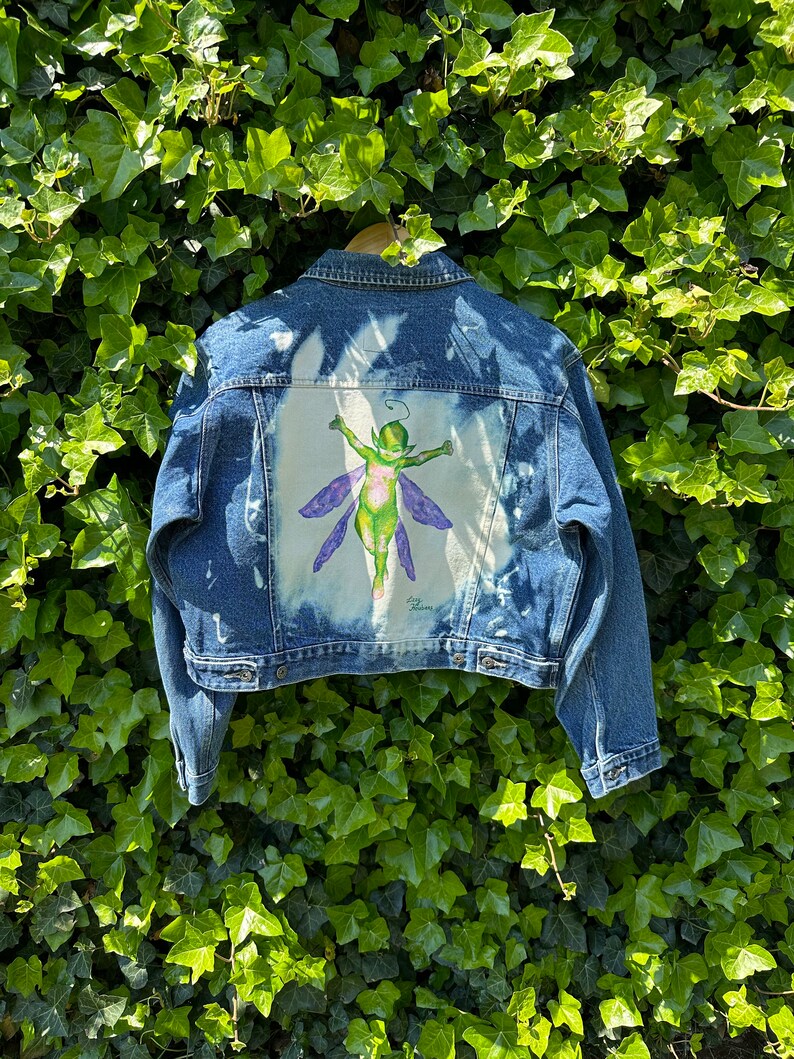 Pu&ograve; includere: Una giacca di jeans blu con un design sbiancato che presenta una fata verde con ali viola sul retro. La fata &egrave; dipinta in uno stile fantasioso.