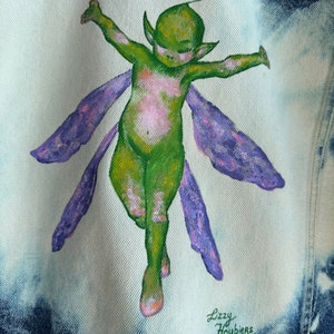 Pu&ograve; includere: Una giacca di jeans con una fata verde dipinta a mano con ali viola. La fata &egrave; raffigurata con le braccia distese e un'espressione fantasiosa. La firma dell'artista, "Lizzy Houbiers", &egrave; visibile nell'angolo in basso a destra.