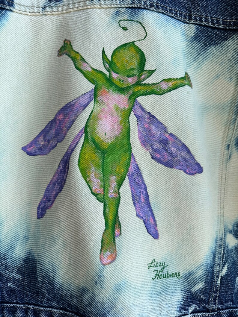 Pu&ograve; includere: Una giacca di jeans con una fata verde dipinta a mano con ali viola. La fata &egrave; raffigurata con le braccia distese e un'espressione fantasiosa. La firma dell'artista, "Lizzy Houbiers", &egrave; visibile nell'angolo in basso a destra.