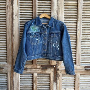 Pu&ograve; includere: Una giacca di jeans blu con schizzi di vernice bianca e un disegno floreale verde sul lato sinistro del petto. La giacca &egrave; appesa a un appendiabiti di legno su uno sfondo di legno invecchiato.