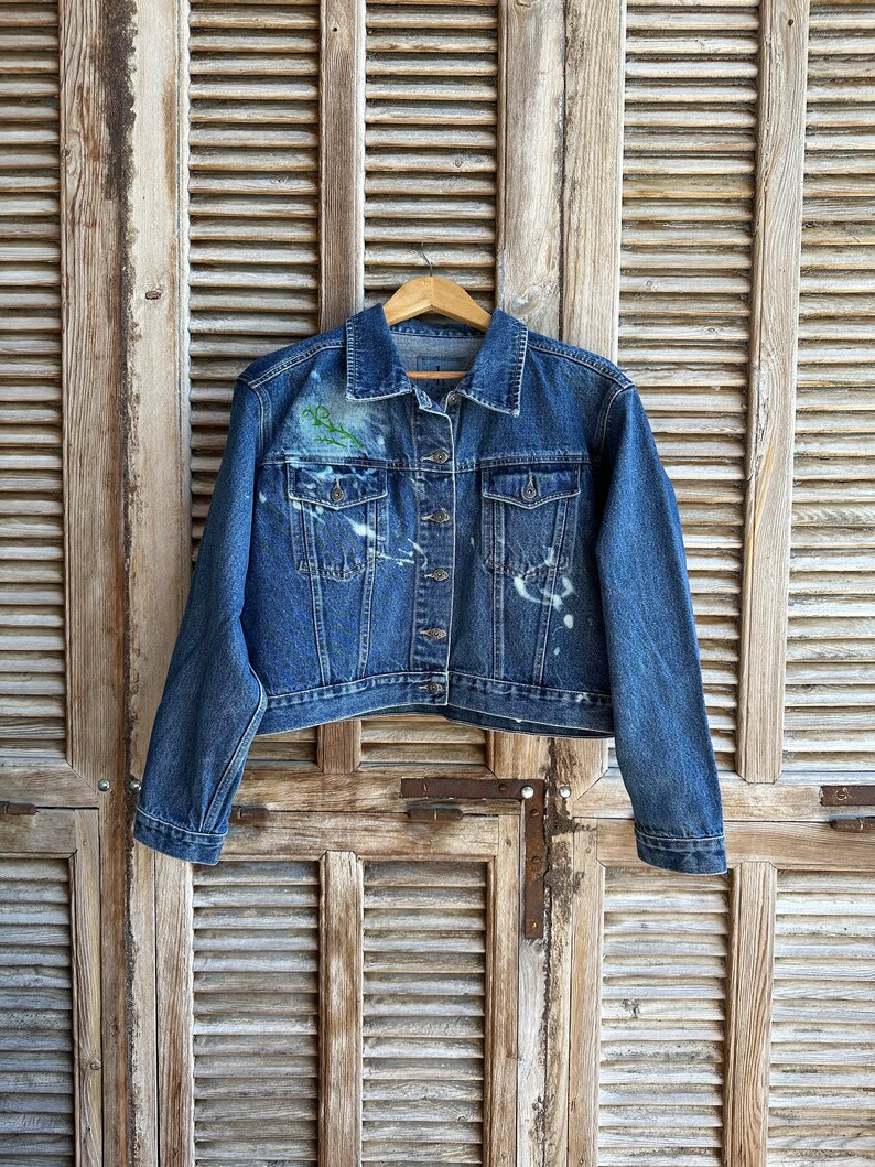 Pu&ograve; includere: Una giacca di jeans blu con schizzi di vernice bianca e un disegno floreale verde sul lato sinistro del petto. La giacca &egrave; appesa a un appendiabiti di legno su uno sfondo di legno invecchiato.