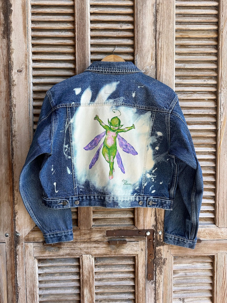 Pu&ograve; includere: Una giacca di jeans blu con un design sbiancato sul retro. Il design presenta una fata verde con ali viola. La firma dell'artista, "Lizzy Meadows", &egrave; visibile sotto la fata.