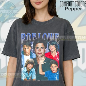 Puede incluir: Una camiseta Comfort Colors gris oscuro con un gráfico de Rob Lowe y otros actores. El texto "ROB LOWE" está en azul, arqueado en la parte superior. La camiseta tiene un aspecto vintage y desgastado.