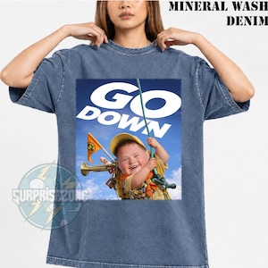 Op de afbeelding: Een t-shirt van mineraal gewassen denim met een grafische print van een kind in scoutuniform dat een touw vasthoudt tegen een blauwe lucht. De tekst GO DOWN staat in grote witte letters boven het kind gedrukt.