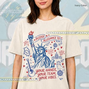 May include: An ivory-coloured t-shirt featuring a blue and red graphic print of the Statue of Liberty, a guitar, a cat, and the text "PLEDGE ALLEGIANCE TO YOUR HANDS, YOUR TEAM, YOUR VIBES." The design includes stars, hearts, fireworks, and a disco ball.