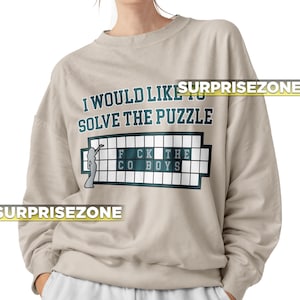 Könnte beinhalten: Ein hellbeiger Pullover mit dem Text "I would like to solve the puzzle" und einem Spielbrett mit dem Text "FCK THE CO BOYS".