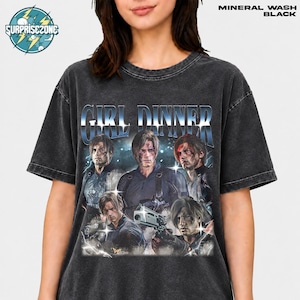 May include: A black mineral wash t-shirt featuring the text "GIRL DINNER" in a metallic blue font. The graphic displays multiple depictions of a male character with brown hair, one holding a handgun, set against a dark, stormy background.