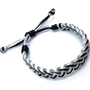 May include: A braided bracelet crafted from alternating strands of black and silver-white cord. The design features a circular braided band with two loose, adjustable cord ends, each finished with a small knot at the tip to secure the bracelet.