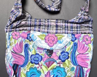 Puede incluir: Un bolso de hombro estilo hobo con tela tejida a cuadros azules y morados en la parte superior e inferior. El panel central es de tela blanca decorada con bordados florales vibrantes y dos grandes figuras de aves rosas y azules con broche.