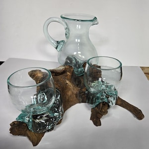 May include: A clear glass jug with a handle and two clear glass cups are mounted on a piece of driftwood. The cups and jug have a teal-coloured base. The driftwood is brown.