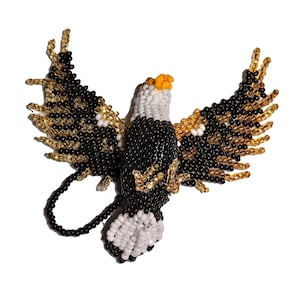 May include: A handcrafted beaded eagle brooch featuring a spread-wing design. The bird is constructed from small black, white, gold, and orange glass beads, depicting a bald eagle in flight with its talons extended and a circular beaded loop.