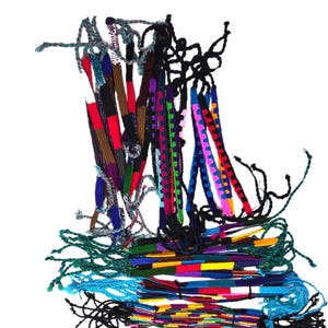 May include: A collection of woven friendship bracelets featuring various geometric patterns and a vibrant color palette including red, blue, green, yellow, purple, and black. The bracelets are made of thin thread with loose ends for tying.