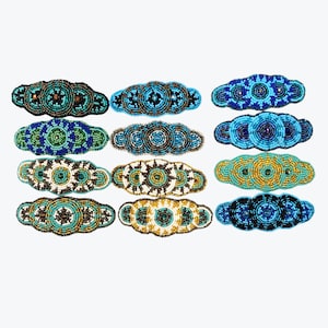 May include: A collection of twelve rectangular hair barrettes arranged in a grid, each featuring intricate beadwork in various circular patterns. The beads come in shades of blue, green, white, gold, and black, creating distinct geometric designs.