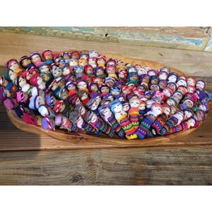 May include: A large collection of small, handcrafted worry dolls arranged in a wooden bowl. Each doll features a painted face and is wrapped in vibrant, multi-colored fabric strips. The dolls are densely packed together, displaying a variety of bright textile patterns including stripes and solid colors, creating a dense, colorful display of miniature figures.
