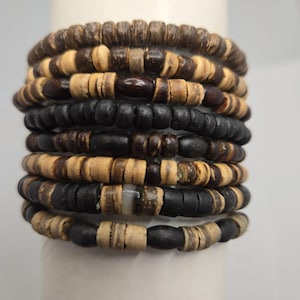 May include: A stack of seven bracelets made from small, flat, circular beads in alternating shades of dark brown, light tan, and matte black. The beads are strung together, creating a textured, layered appearance with varied bead shapes.