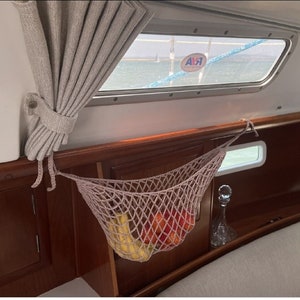 May include: A beige net hammock hanging from the ceiling of a boat, filled with bananas and red apples. The hammock is made of a woven material and is hanging from a wooden beam. The boat has a wooden interior and a window with a blue sky and water visible outside.