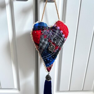 May include: A heart-shaped fabric ornament with a patchwork design in red, blue, and grey plaid patterns. It features a cream-coloured lace trim, a silver charm, and a navy blue tassel. The ornament hangs from a gold cord.