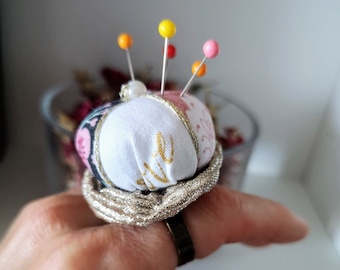 Può includere: Un cuscino per spilli bianco con una scritta dorata che dice "cute" e una varietà di spilli colorati. Il cuscino per spilli è avvolto in un tessuto argentato e si trova su un dito.