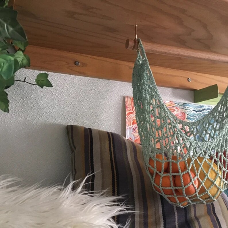 May include: A green crocheted hammock hanging from a wooden beam with a hook. The hammock is filled with oranges and a yellow lemon.