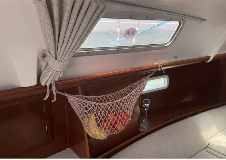 May include: A beige net hammock hanging from the ceiling of a boat, filled with bananas and red apples. The hammock is made of a woven material and is hanging from a wooden beam. The boat has a wooden interior and a window with a blue sky and water visible outside.