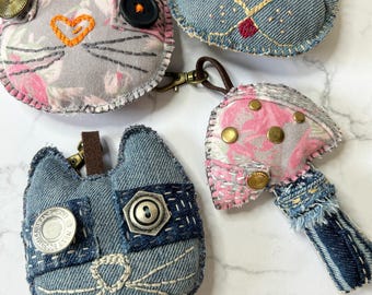 May include: Four handmade fabric keychains featuring animal faces. The keychains are made from denim and floral fabric. The keychains are decorated with buttons, stitching, and embroidery.