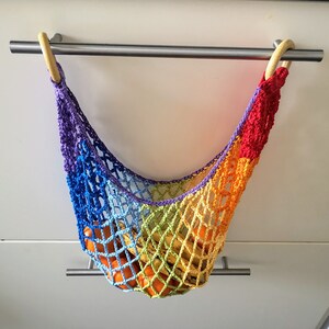 May include: A rainbow crocheted hanging fruit hammock with wooden rings. The hammock is filled with oranges and bananas.