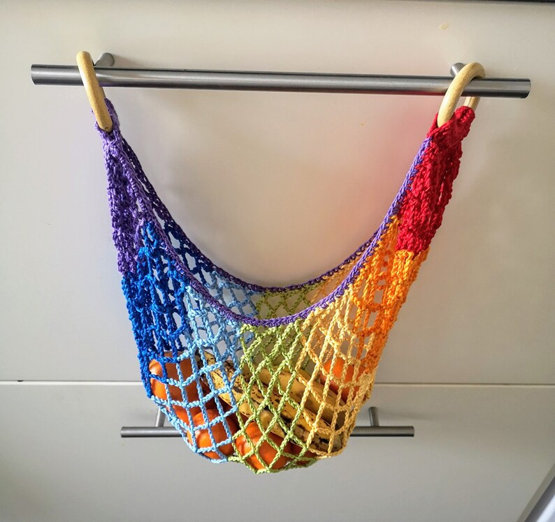 May include: A rainbow crocheted hanging fruit hammock with wooden rings. The hammock is filled with oranges and bananas.