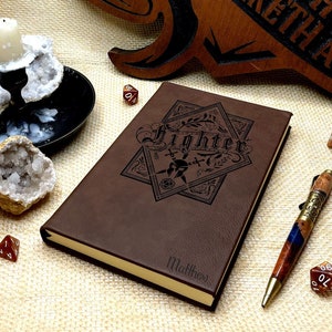 May include: Brown leather journal with a gold-colored edge and a black engraved design of a sword and dice with the word "Fighter" above it. The journal is personalized with the name "Matthew".