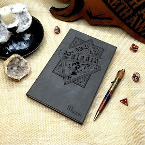 May include: A gray leather-bound journal with the words "Paladin" and "Mason" embossed on the cover. The journal is surrounded by dice and a pen.
