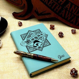 May include: A teal blue leather-bound journal with a black and white illustration of a star with a stylized tree and the word "Runes" inside. The journal has the name "Gina" written on the front cover. A pen with a brass tip and a wooden handle is lying on top of the journal.