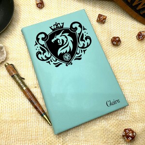 May include: A teal leather journal with a black dragon and a dice design. The journal has the name "Claire" embossed on the front.