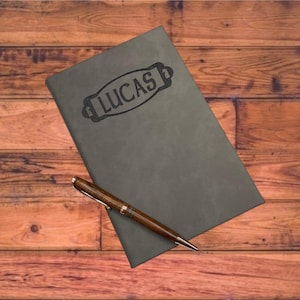 May include: A gray leather-bound journal with the name "LUCAS" embossed on the front cover. A brown pen is lying on top of the journal.