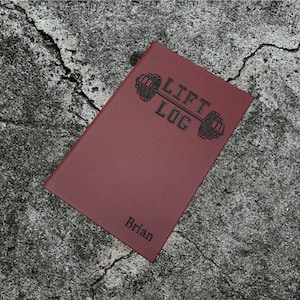 May include: A burgundy leather-bound journal with the text "LIFT LOG" and a graphic of a barbell with weights. The name "Brian" is embossed on the front cover.