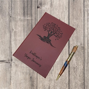 May include: A burgundy leather journal with a tree and a person in a yoga pose engraved on the front. The text "Kalliope's Yoga Journey" is also engraved on the front. A wooden pen is lying next to the journal.