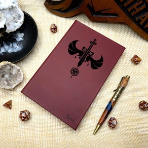 May include: A burgundy leather journal with a black design of a sword with wings and a dice. The journal has the word "Tarvok" embossed on the front.