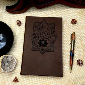 May include: A brown leather-bound journal with the word "Artificer" embossed on the front cover. The journal is surrounded by dice and a pen.