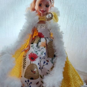Op de afbeelding: Een pop die een gele gebreide cape draagt met witte bontkraag, een witte kant-gezoomde jurk met een bijenkorf en bloemenpatroon, en een gouden kroon met een parel.