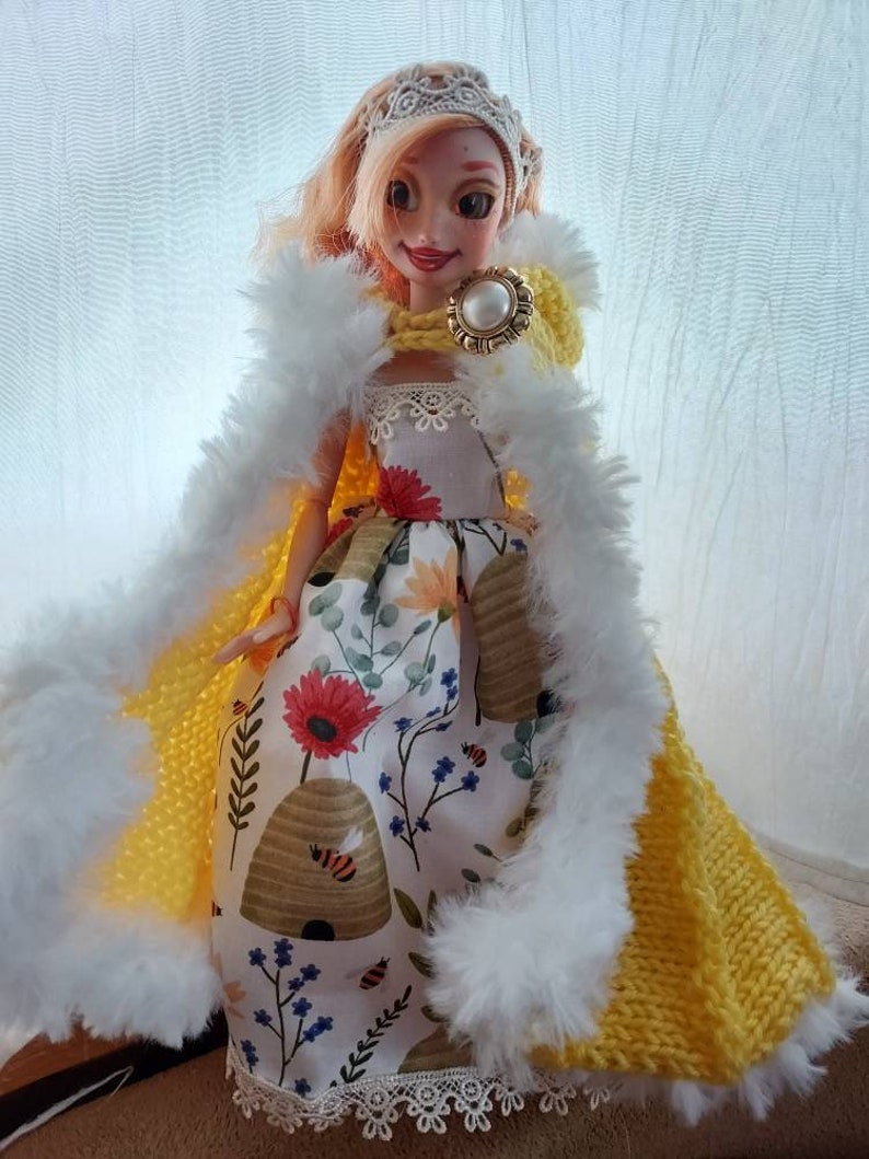 Op de afbeelding: Een pop die een gele gebreide cape draagt met witte bontkraag, een witte kant-gezoomde jurk met een bijenkorf en bloemenpatroon, en een gouden kroon met een parel.