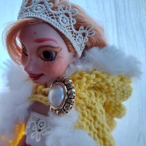 Op de afbeelding: Een pop met blond haar die een witte kanten kroon, een gele gebreide trui met capuchon en een witte bontkraag draagt. De pop heeft een parel- en gouden oorring.