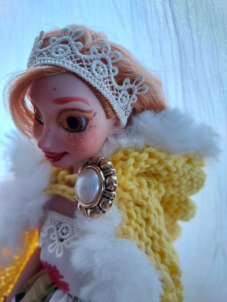Op de afbeelding: Een pop met blond haar die een witte kanten kroon, een gele gebreide trui met capuchon en een witte bontkraag draagt. De pop heeft een parel- en gouden oorring.