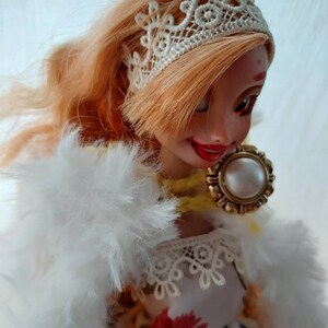 Op de afbeelding: Een pop met lang, blond haar die een wit kant haarbandje, een witte bontjas en een bloemenjurk met een witte kanten rand draagt. De pop draagt een gouden ketting met een parel hanger.