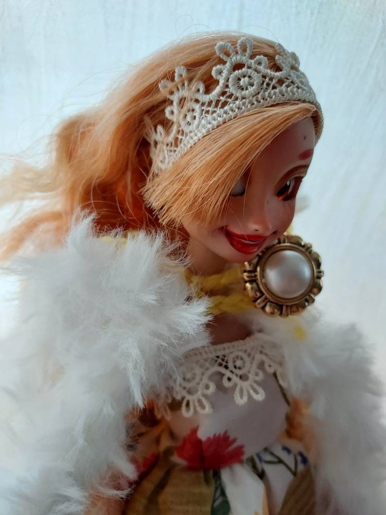 Op de afbeelding: Een pop met lang, blond haar die een wit kant haarbandje, een witte bontjas en een bloemenjurk met een witte kanten rand draagt. De pop draagt een gouden ketting met een parel hanger.