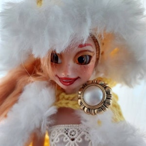 Op de afbeelding: Een pop met blond haar die een gele gehaakte hoed draagt met een witte bontkraag, een witte bont sjaal en een witte kanten jurk. De pop houdt een gouden broche vast met een parel in het midden.