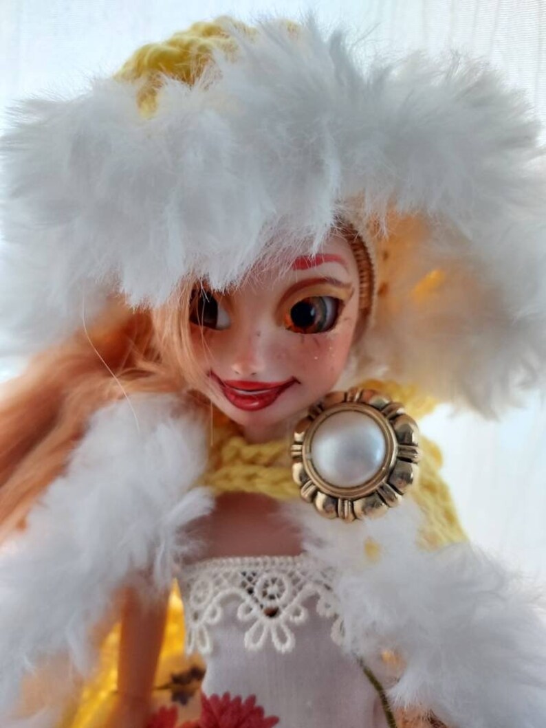 Op de afbeelding: Een pop met blond haar die een gele gehaakte hoed draagt met een witte bontkraag, een witte bont sjaal en een witte kanten jurk. De pop houdt een gouden broche vast met een parel in het midden.