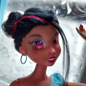 May include: A black haired doll with pink highlights in her hair. She is wearing a blue and white dress and has blue hoop earrings. Her eyes are painted with a blue and red star design.