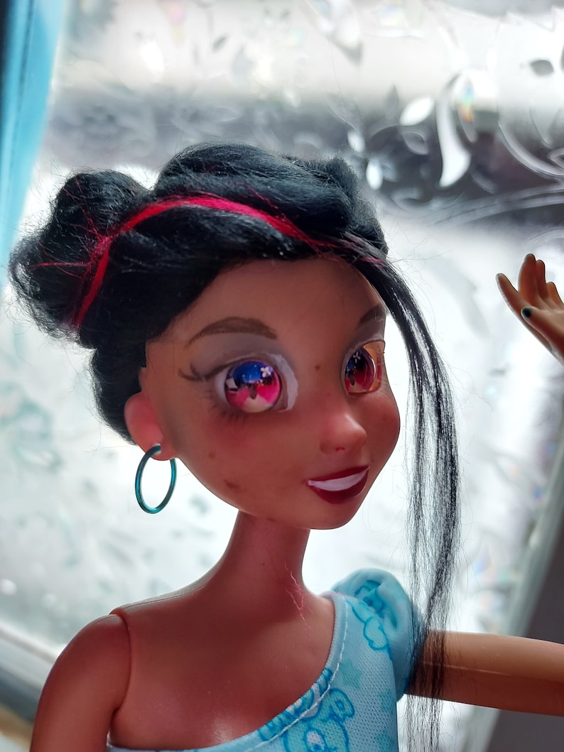May include: A black haired doll with pink highlights in her hair. She is wearing a blue and white dress and has blue hoop earrings. Her eyes are painted with a blue and red star design.